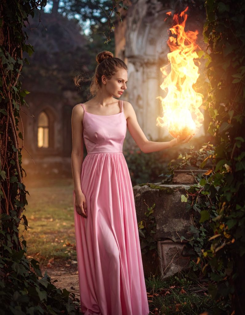A young woman in a long pink dress standing in ivy-covered ruins, calmly holding a large, bright ball of fire in her hand