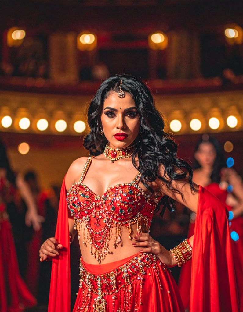 A female belly dancer in a red and gold beaded costume standing in front of warm bokeh lights.